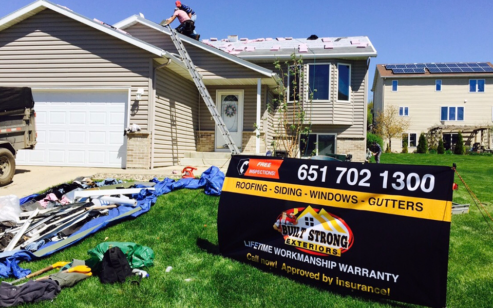Built Strong Exteriors' team members working on roof repair project. In front of the house there's a promotional sign that states Built Strong Exteriors' business info.